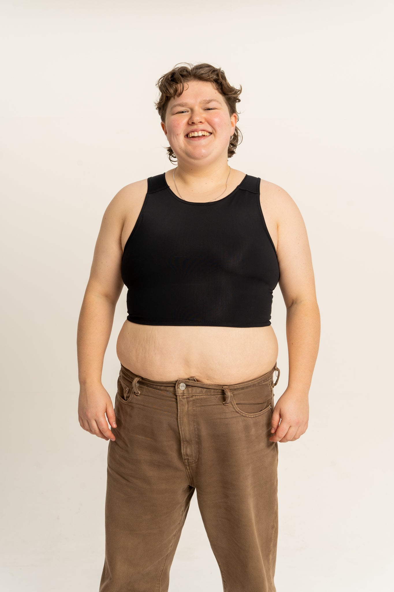Person wearing a black short chest binder and brown trousers against a white background