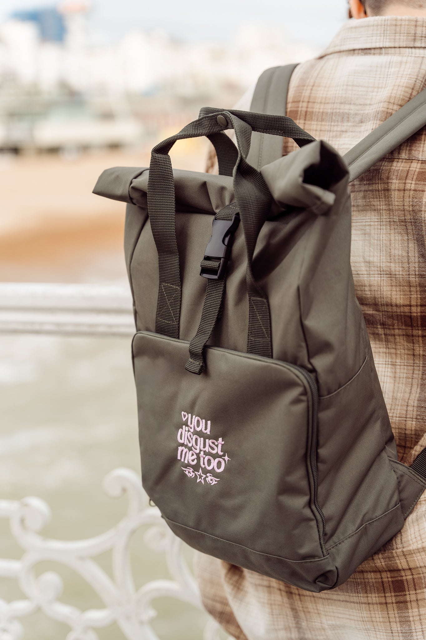 Person wearing a green backpack with pink embroidery reading 'You disgust me too' text, standing outdoors.