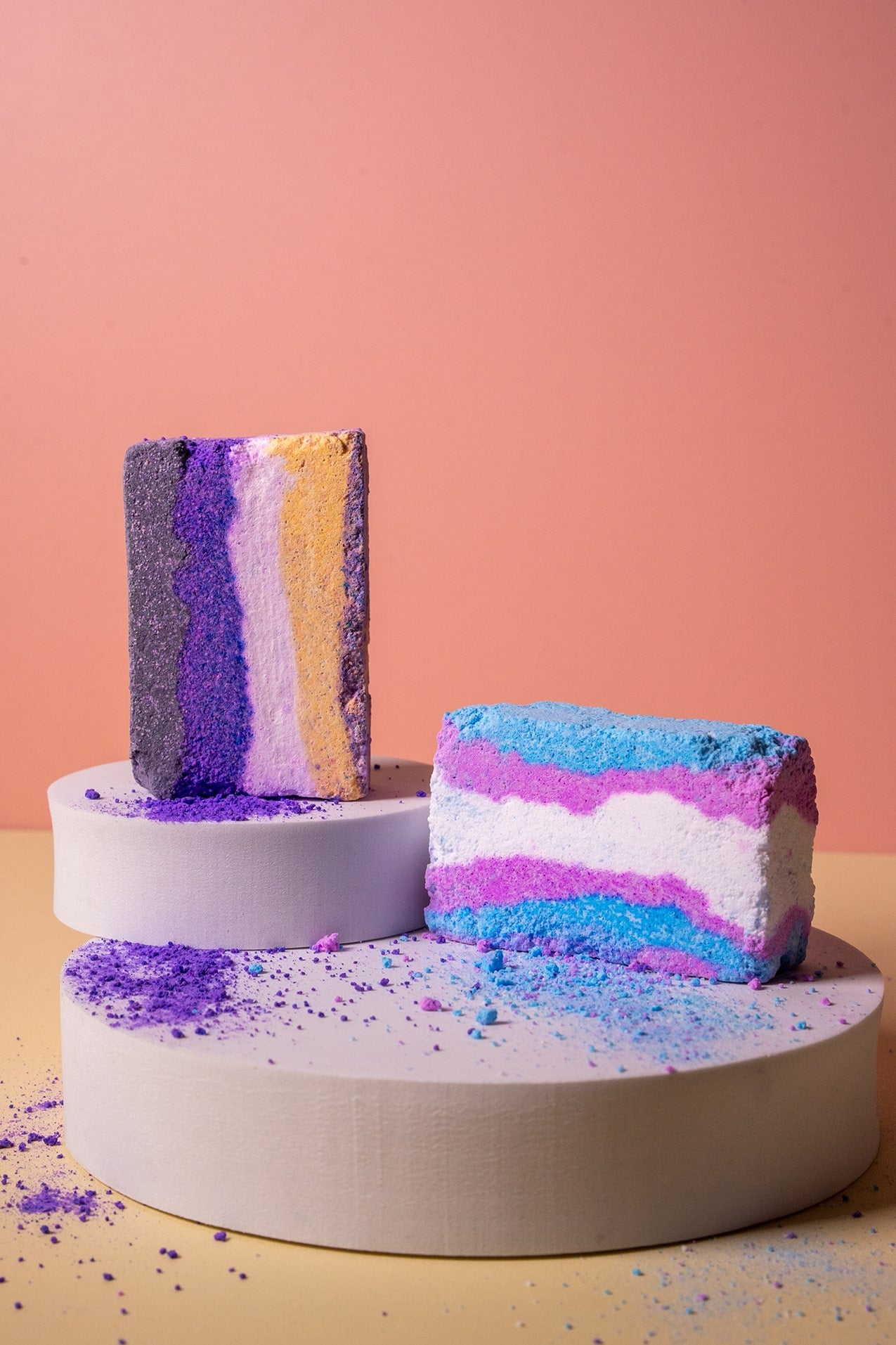 Colorful trans and non binary flag bath bombs on a white stand against a pink background