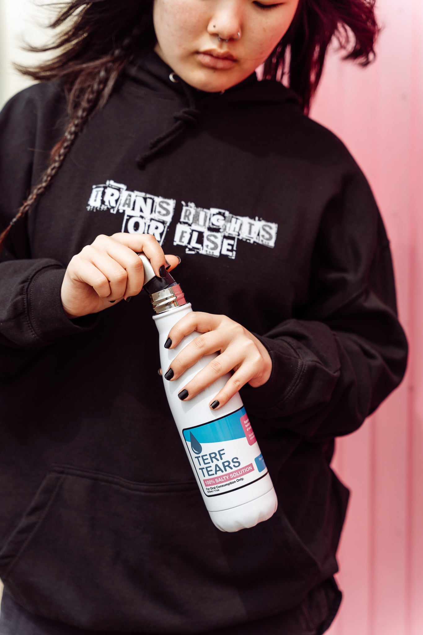 Person wearing a black hoodie with text, holding a white bottle against a pink background